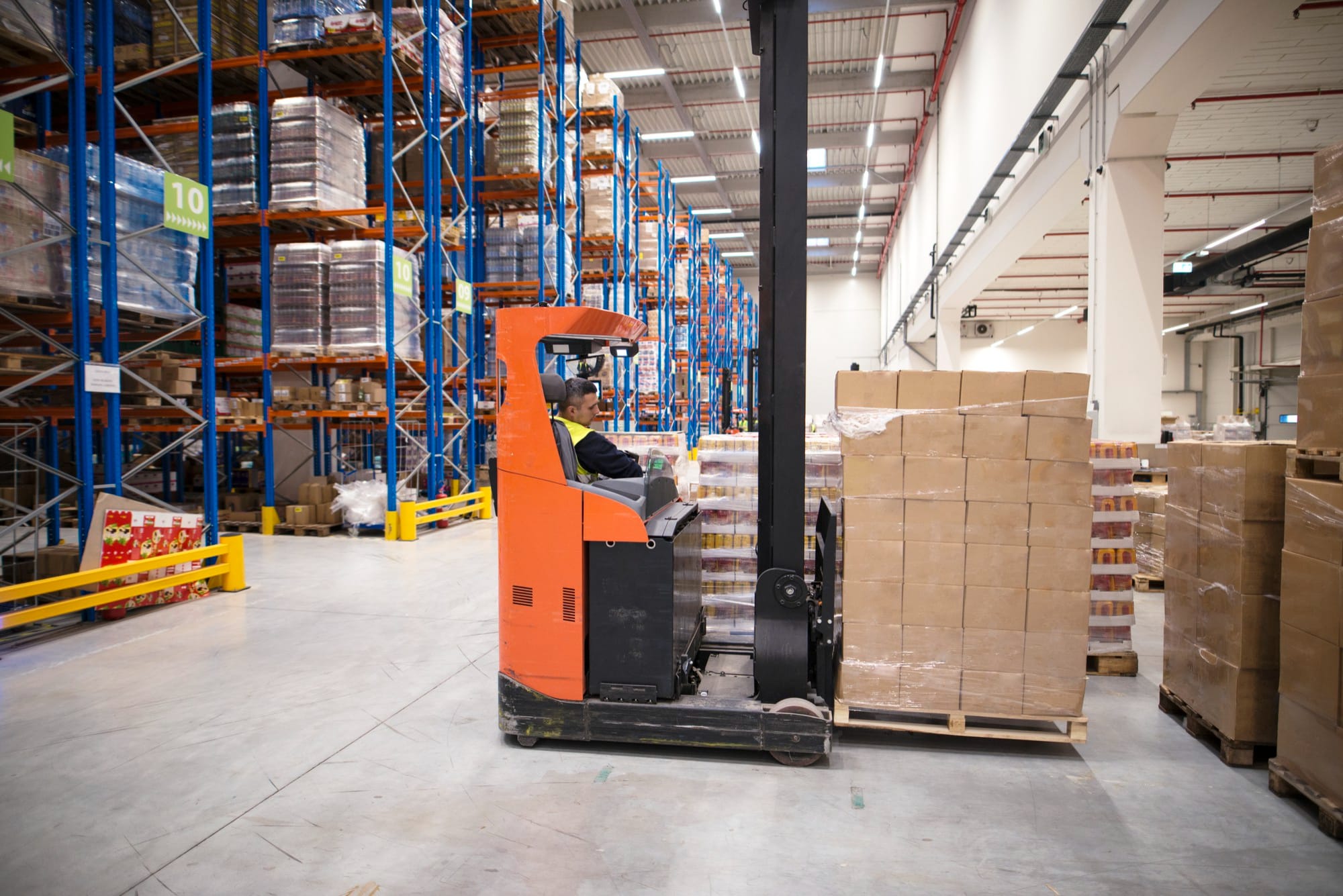 Forklift operator moving palletised stock through a warehouse aisle lined with high-bay pallet racking