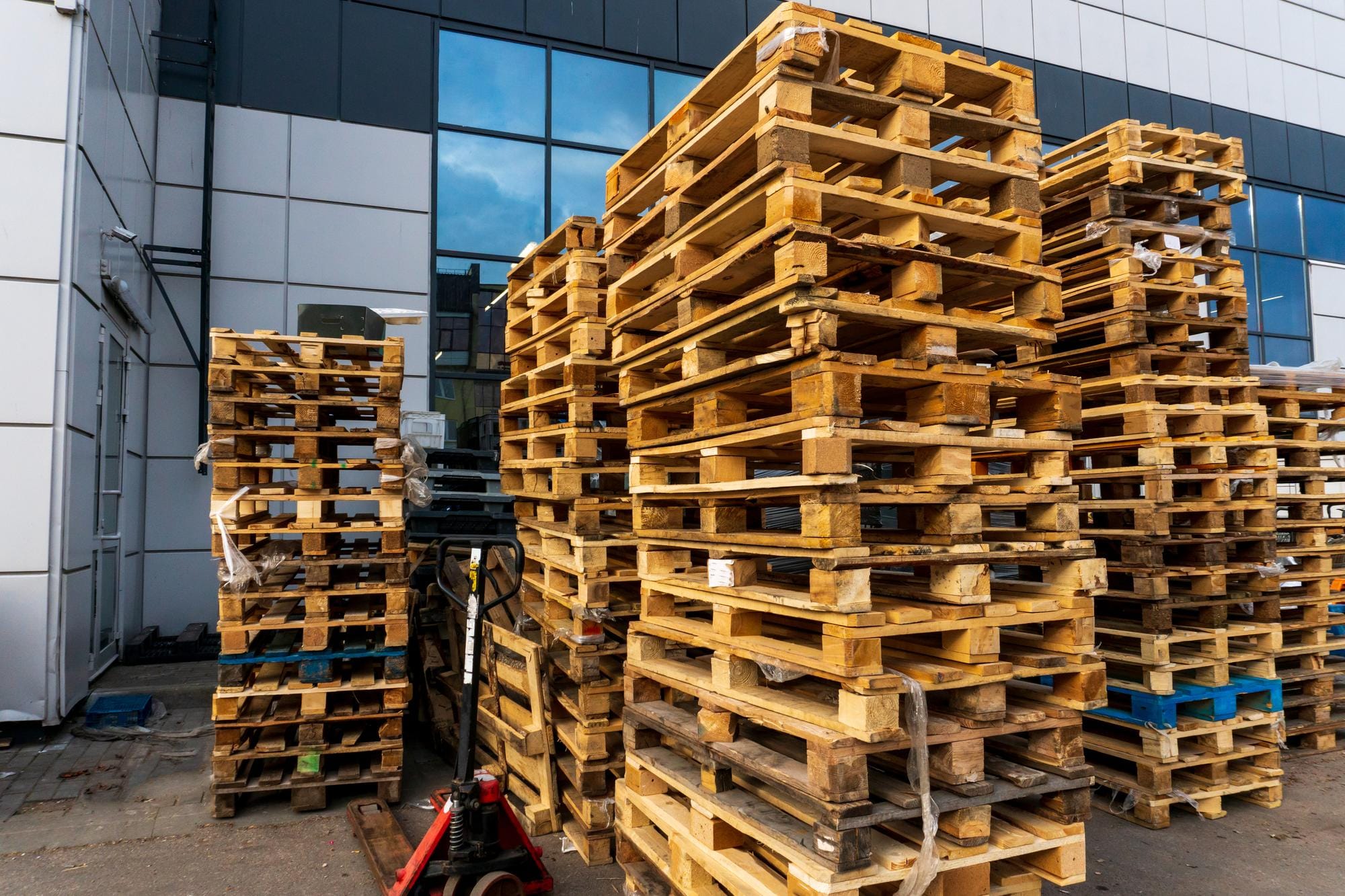 Large stacks of wooden pallets stored outdoors with a modern warehouse building and front windows in the background.