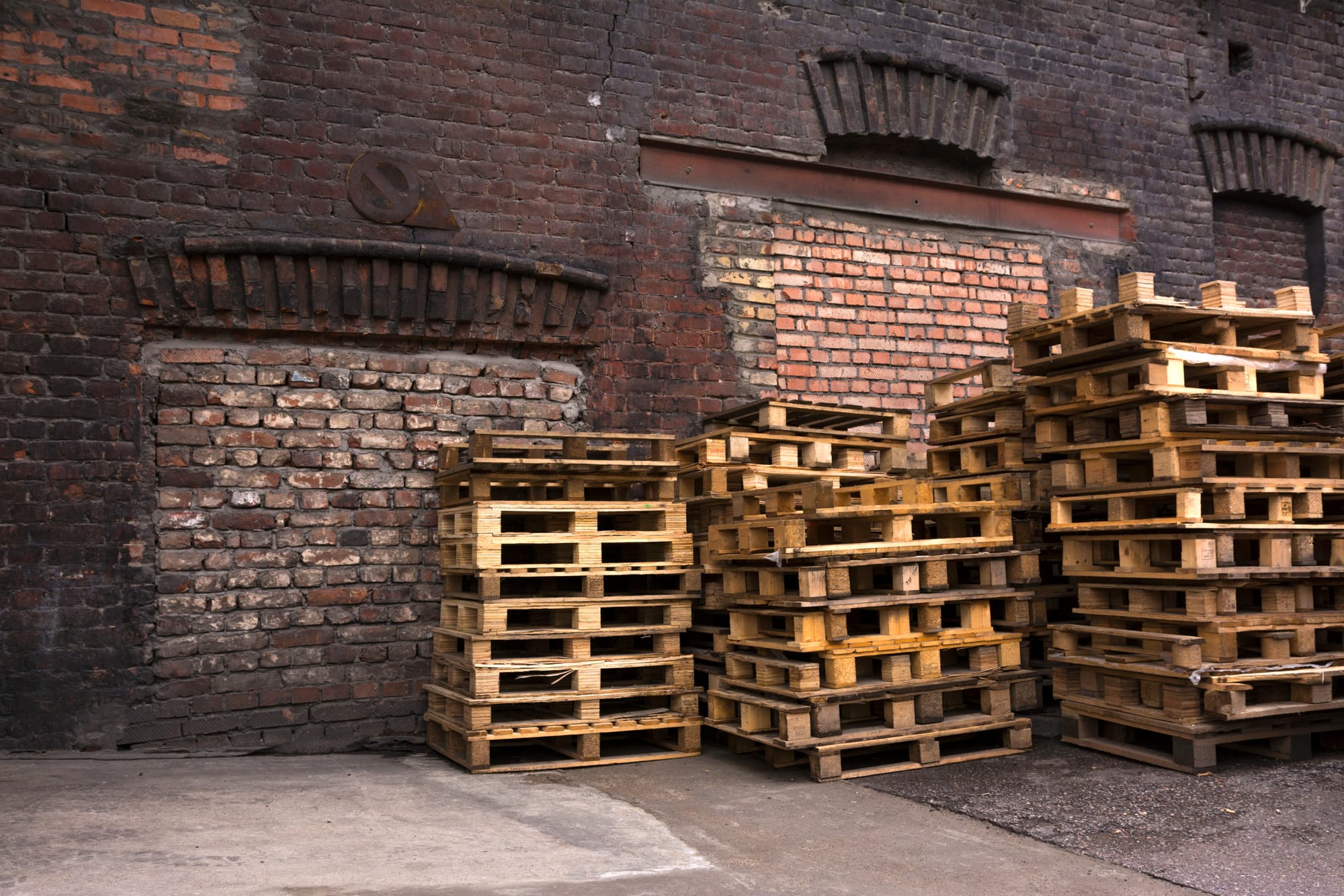 Multiple stacks of wooden pallets neatly arranged in a yard with a brick warehouse building in the background.