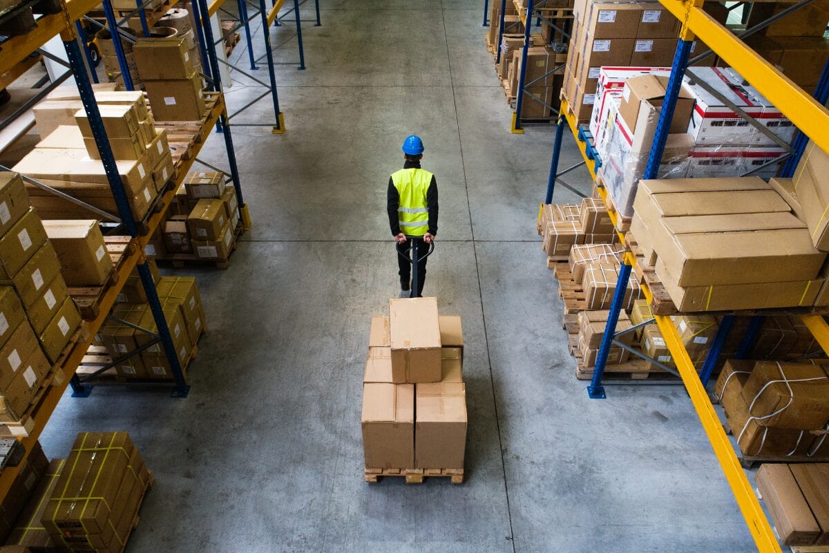 Warehouse worker using a pallet jack to move boxed goods on a pallet between storage racks